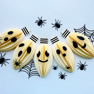 May include: A string of six white paper lanterns with black cut-out faces, decorated with black paper spiders and spiderwebs. The lanterns have different facial expressions, including a smiling face, a frowning face, and a surprised face.