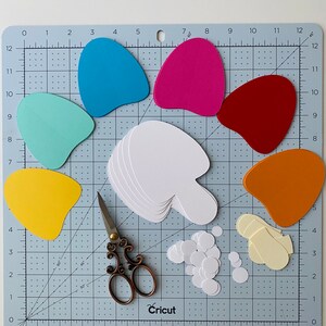 May include: Blue, green, pink, red, yellow and orange paper shapes on a cutting mat with a pair of scissors, a stack of white paper shapes, and a pile of white circles.