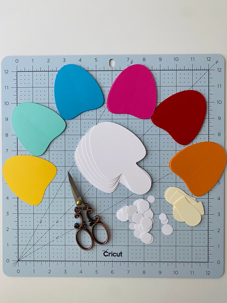 May include: Blue, green, pink, red, yellow and orange paper shapes on a cutting mat with a pair of scissors, a stack of white paper shapes, and a pile of white circles.