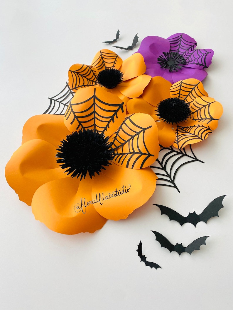 K&ouml;nnte beinhalten: Orangefarbene und violette Papierblumen mit schwarzen Spinnweben und Flederm&auml;usen. Die Blumen sind &uuml;bereinander gelegt und haben einen 3D-Effekt. Die Flederm&auml;use sind aus schwarzem Papier ausgeschnitten und fliegen in der Luft. Die Blumen sind mit schwarzen Pompons in der Mitte verziert.