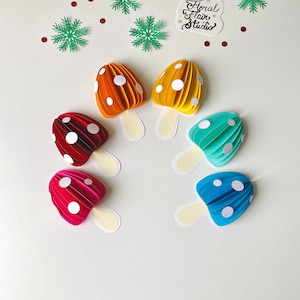 May include: Six paper mushroom decorations in various colours, including red, pink, yellow, orange, blue, and green. Each mushroom has white polka dots and a white stem. The mushrooms are arranged in a semi-circle on a white background. The text "Floral Flair Studio" is visible in the top right corner of the image.