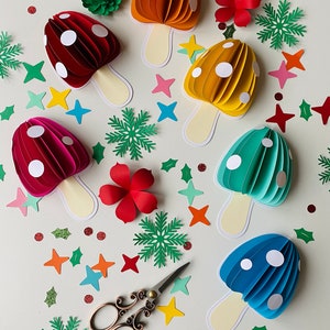 May include: A collection of six papercraft mushrooms in various colours, including red, orange, yellow, green, and blue. Each mushroom has white polka dots and a white stem. The mushrooms are arranged on a white surface with various paper cutouts, including stars, snowflakes, and leaves.