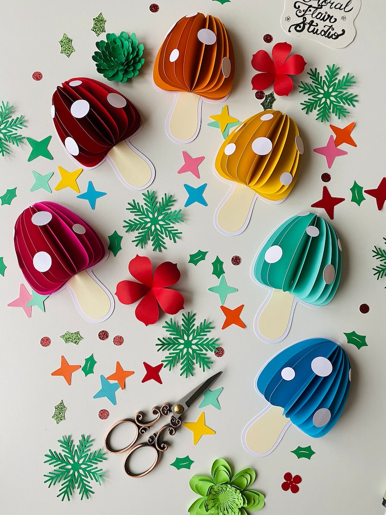 May include: A collection of six papercraft mushrooms in various colours, including red, orange, yellow, green, and blue. Each mushroom has white polka dots and a white stem. The mushrooms are arranged on a white surface with various paper cutouts, including stars, snowflakes, and leaves.