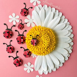 Pode incluir: Obra de arte em papel com uma margarida branca com um centro amarelo cercada por joaninhas vermelhas sobre um fundo rosa. A margarida tem múltiplas pétalas brancas e as joaninhas têm manchas pretas e corações nas suas asas. A obra é assinada "@floralflairstudio".