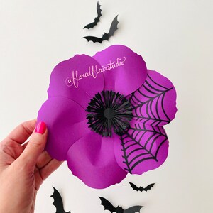 K&ouml;nnte beinhalten: Eine lila Papierblume mit einem schwarzen Spinnennetz-Design und einer schwarzen Papiermitte. Die Blume ist von schwarzen Papierflederm&auml;usen umgeben.