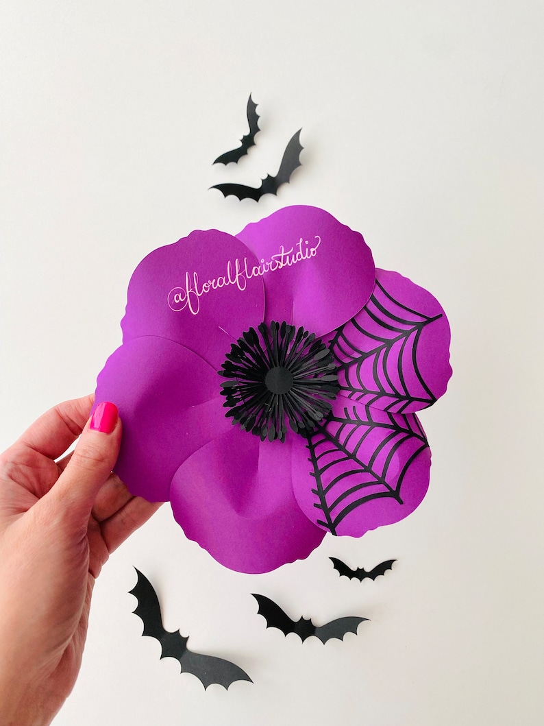 K&ouml;nnte beinhalten: Eine lila Papierblume mit einem schwarzen Spinnennetz-Design und einer schwarzen Papiermitte. Die Blume ist von schwarzen Papierflederm&auml;usen umgeben.