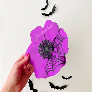 K&ouml;nnte beinhalten: Eine lila Papierblume mit einem schwarzen Spinnennetz-Design und einer schwarzen Mitte. Die Blume wird in einer Hand vor einem wei&szlig;en Hintergrund gehalten. Es sind mehrere schwarze Papierflederm&auml;use um die Blume herum verstreut.