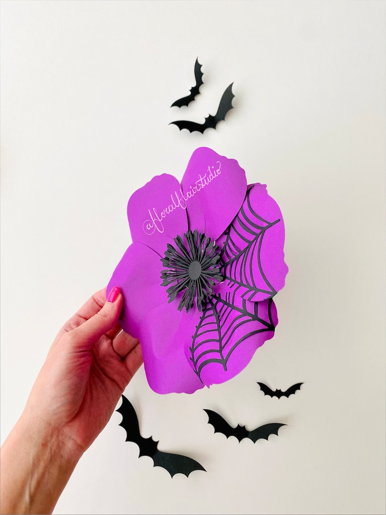 K&ouml;nnte beinhalten: Eine lila Papierblume mit einem schwarzen Spinnennetz-Design und einer schwarzen Mitte. Die Blume wird in einer Hand vor einem wei&szlig;en Hintergrund gehalten. Es sind mehrere schwarze Papierflederm&auml;use um die Blume herum verstreut.