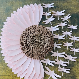 Peut inclure: Un tournesol en papier artisanal avec des pétales roses et un centre brun, entouré d'un groupe de libellules en papier blanc sur un fond en bois. Le texte "@floralflairstudio" est en bas de l'image.