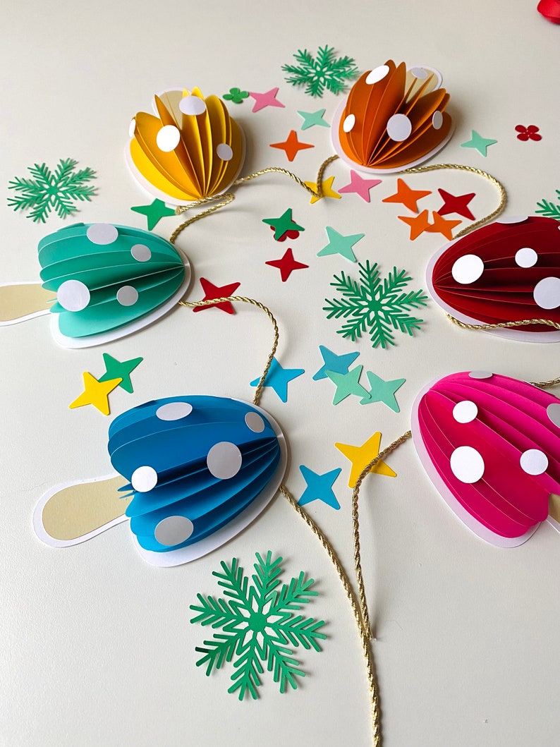 May include: A colourful paper mushroom garland with a gold string. The mushrooms are in shades of red, pink, blue, green, and yellow. Each mushroom has white polka dots. The garland is surrounded by green snowflakes and yellow stars.