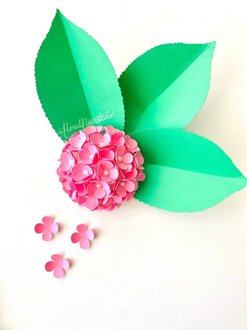 Peut inclure: Une fleur en papier rose avec un centre blanc, entour&eacute;e de trois feuilles en papier vert. La fleur est compos&eacute;e de nombreuses petites p&eacute;tales en papier et a un aspect textur&eacute;. Les feuilles sont &eacute;galement textur&eacute;es et ont un bord festonn&eacute;.