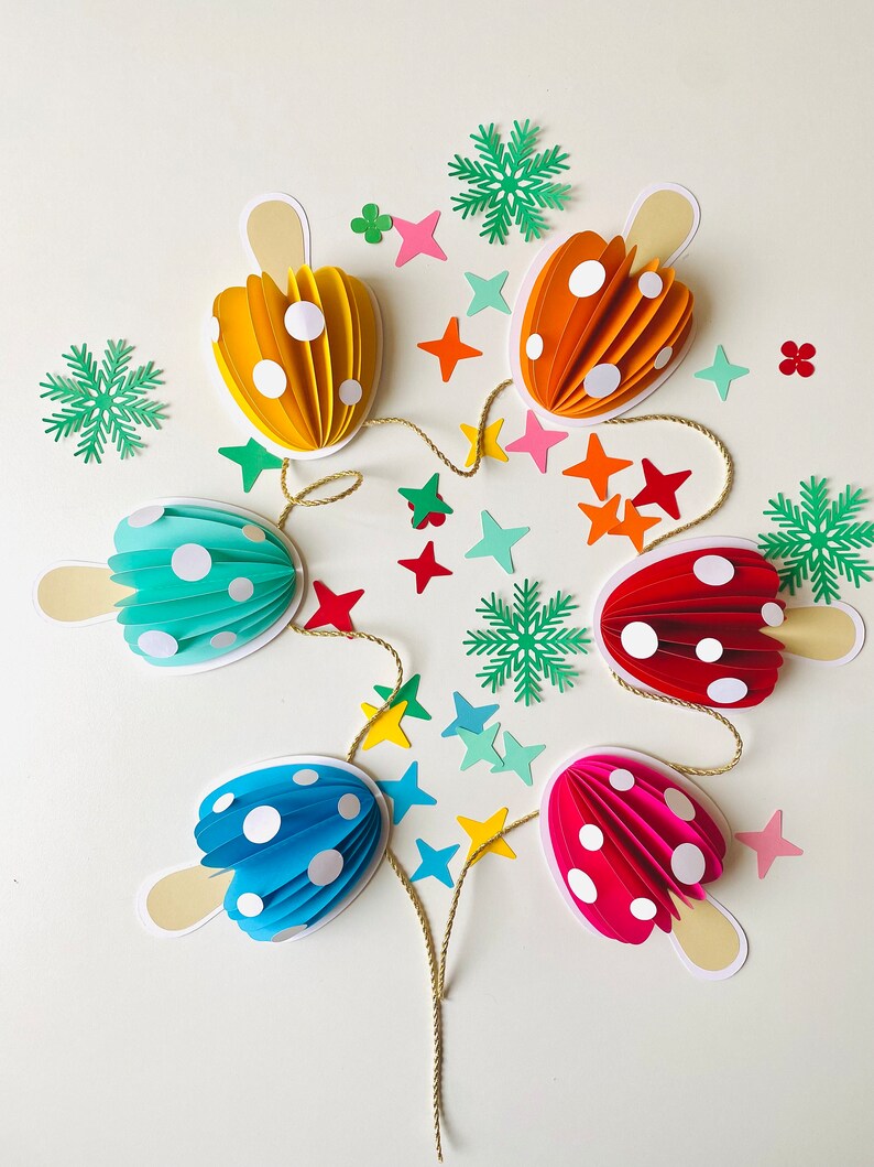 May include: A string of paper mushroom decorations in various colours, including yellow, orange, red, blue, and pink. The mushrooms are decorated with white polka dots and are connected by a gold string. The decorations are surrounded by small paper stars and snowflakes.