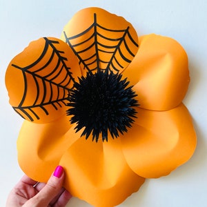 K&ouml;nnte beinhalten: Eine gro&szlig;e orangefarbene Papierblume mit schwarzen Spinnweben und einer schwarzen Papiermitte. Die Blume besteht aus mehreren Papierschichten und hat einen 3D-Effekt.