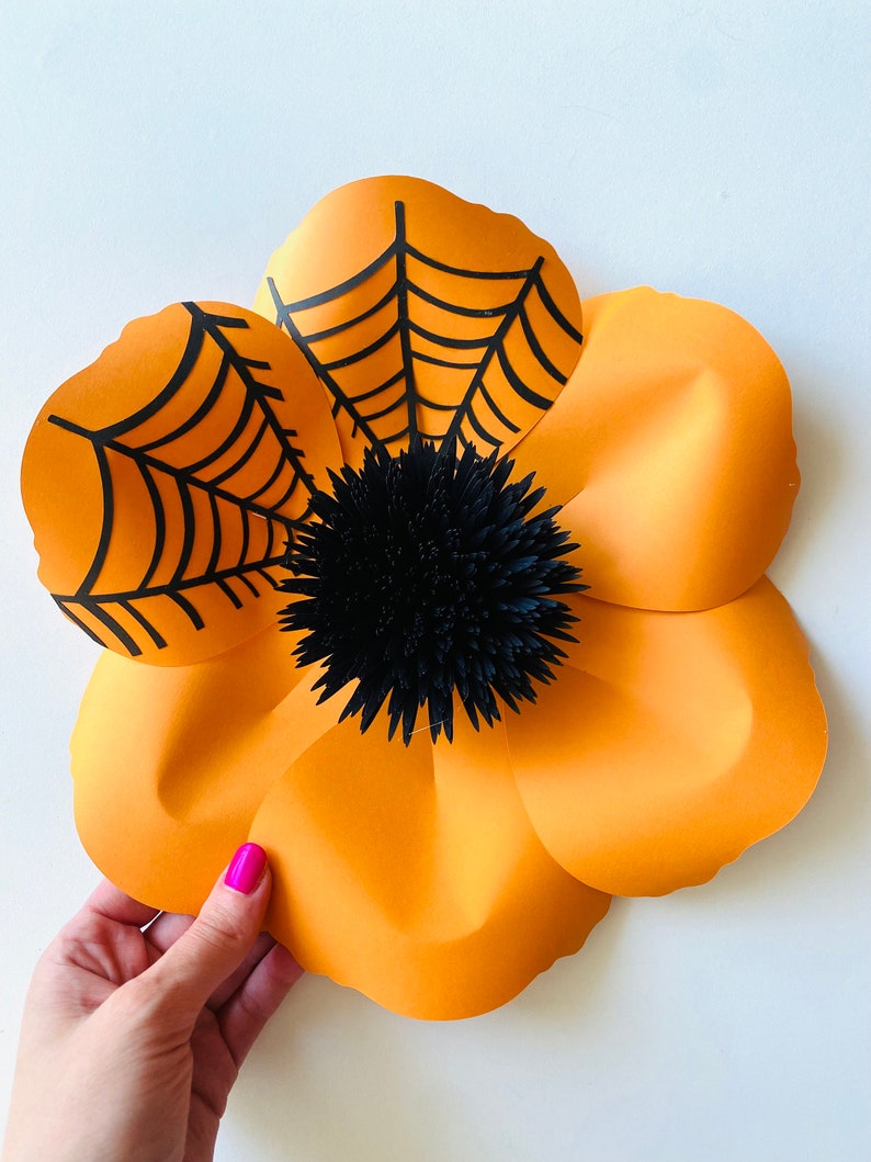 K&ouml;nnte beinhalten: Eine gro&szlig;e orangefarbene Papierblume mit schwarzen Spinnweben und einer schwarzen Papiermitte. Die Blume besteht aus mehreren Papierschichten und hat einen 3D-Effekt.