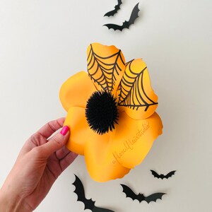 K&ouml;nnte beinhalten: Eine gro&szlig;e orangefarbene Papierblume mit einem schwarzen Spinnennetz-Design und einem schwarzen Pompon-Zentrum. Die Blume ist von schwarzen Papierflederm&auml;usen umgeben.