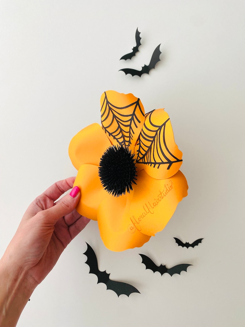 K&ouml;nnte beinhalten: Eine gro&szlig;e orangefarbene Papierblume mit einem schwarzen Spinnennetz-Design und einem schwarzen Pompon-Zentrum. Die Blume ist von schwarzen Papierflederm&auml;usen umgeben.