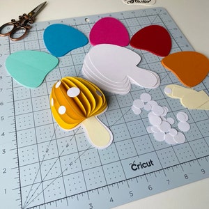 May include: A yellow paper mushroom with white polka dots, a white stem, and white circles on a blue cutting mat. The mushroom is made of multiple layers of paper, creating a 3D effect. There are also other paper shapes in various colours, including red, orange, pink, and blue.