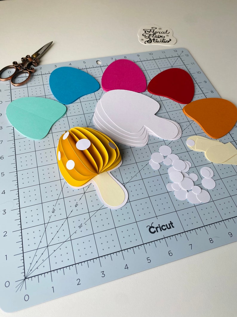 May include: A yellow paper mushroom with white polka dots, a white stem, and white circles on a blue cutting mat. The mushroom is made of multiple layers of paper, creating a 3D effect. There are also other paper shapes in various colours, including red, orange, pink, and blue.