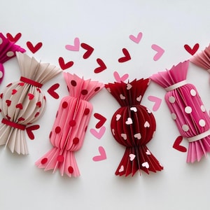 Puede incluir: Seis decoraciones de caramelo de papel en varios tonos de rosa y rojo con lunares blancos o corazones. El caramelo está doblado para crear un efecto 3D.