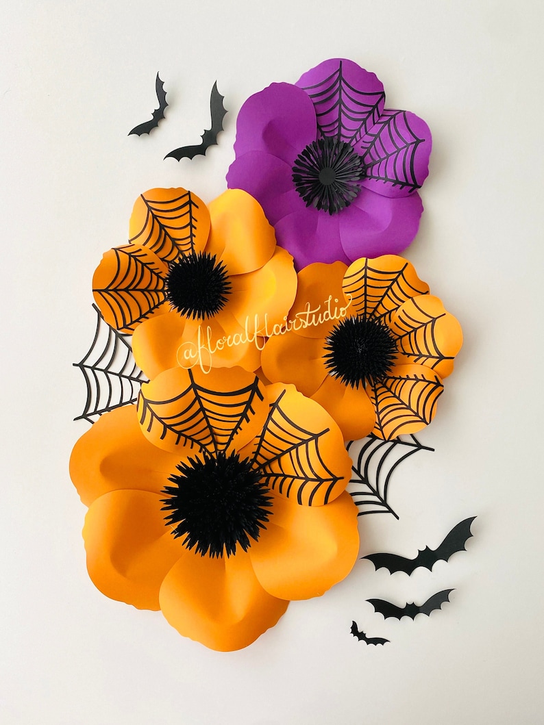 K&ouml;nnte beinhalten: Drei gro&szlig;e Papierblumen mit schwarzen Spinnweben darauf. Die Blumen sind orange und lila. Es gibt auch mehrere schwarze Papierflederm&auml;use auf wei&szlig;em Hintergrund.