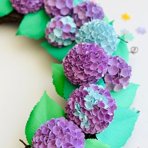 Peut inclure: Une couronne de fleurs en papier avec des fleurs d'hortensias violettes et bleues et des feuilles vertes. La couronne est faite de brindilles et est parfaite pour une d&eacute;coration de printemps ou d'&eacute;t&eacute;.