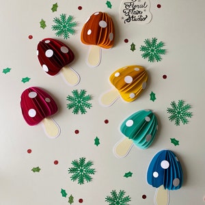 May include: Seven paper mushroom decorations in a variety of colours, including red, orange, yellow, green, and blue. Each mushroom has white polka dots and a cream-coloured stem. The mushrooms are arranged on a white background with green holly leaves and snowflakes.