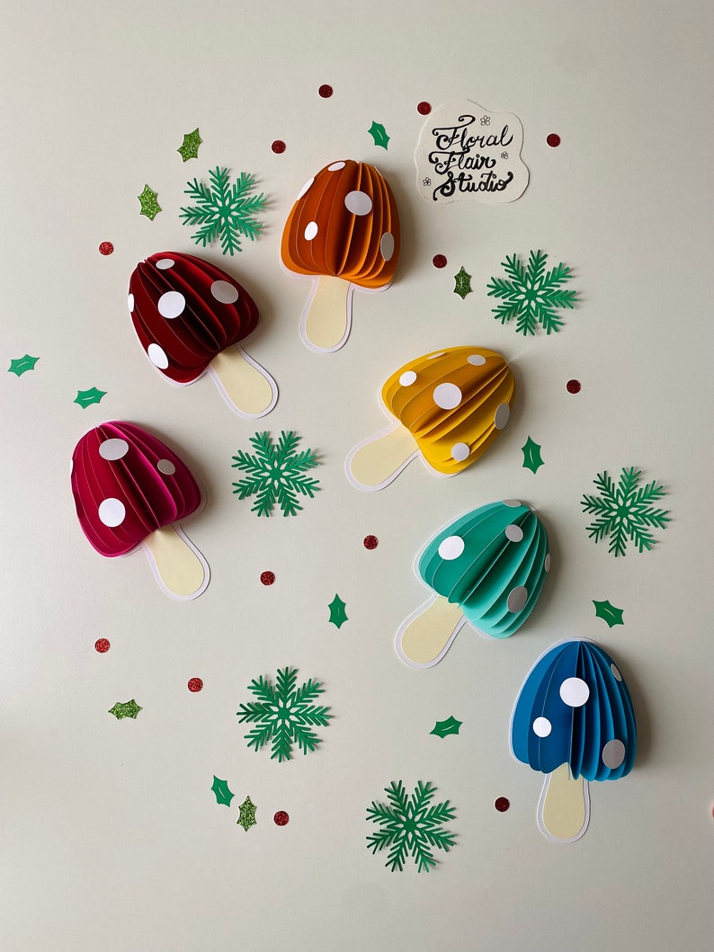 May include: Seven paper mushroom decorations in a variety of colours, including red, orange, yellow, green, and blue. Each mushroom has white polka dots and a cream-coloured stem. The mushrooms are arranged on a white background with green holly leaves and snowflakes.