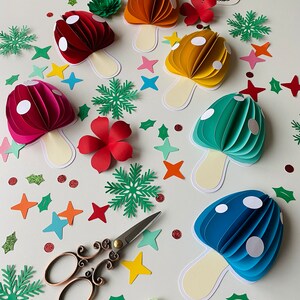 May include: A set of six paper mushroom decorations in various colours, including red, orange, yellow, green, and blue. Each mushroom has white polka dots and a cream-coloured stem. The mushrooms are surrounded by green leaves, red flowers, and white stars.