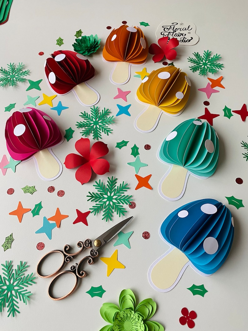 May include: A set of six paper mushroom decorations in various colours, including red, orange, yellow, green, and blue. Each mushroom has white polka dots and a cream-coloured stem. The mushrooms are surrounded by green leaves, red flowers, and white stars.