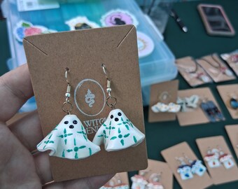 May include: A pair of white ghost earrings with green cross patterns and black eyes. The earrings are displayed on a brown card with a logo. Other earrings are visible in the background.