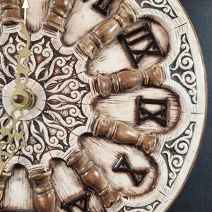 May include: A round wall clock with a cream-colored ceramic face featuring raised, dark brown Roman numerals and decorative spindle-shaped markers. The center displays an intricate, embossed scrollwork pattern, and thin gold-toned metal hands.