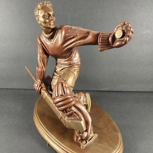 May include: A bronze-toned sculpture of a hockey player in motion, wearing a jersey, gloves, and skates. The figure holds a hockey stick in one hand and extends the other hand forward, catching a small, circular, metallic puck-like object.