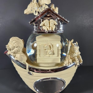 May include: A decorative snow globe featuring a cream-colored ark with a lion, zebra, and giraffe figurines. The globe has a house with sheep inside and a clear glass ball with a family of animals. The base has a silver-colored trim.