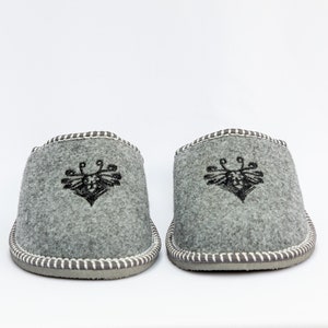May include: A pair of gray wool slippers with a black embroidered bee design. The slippers have a white stitched trim around the edges.