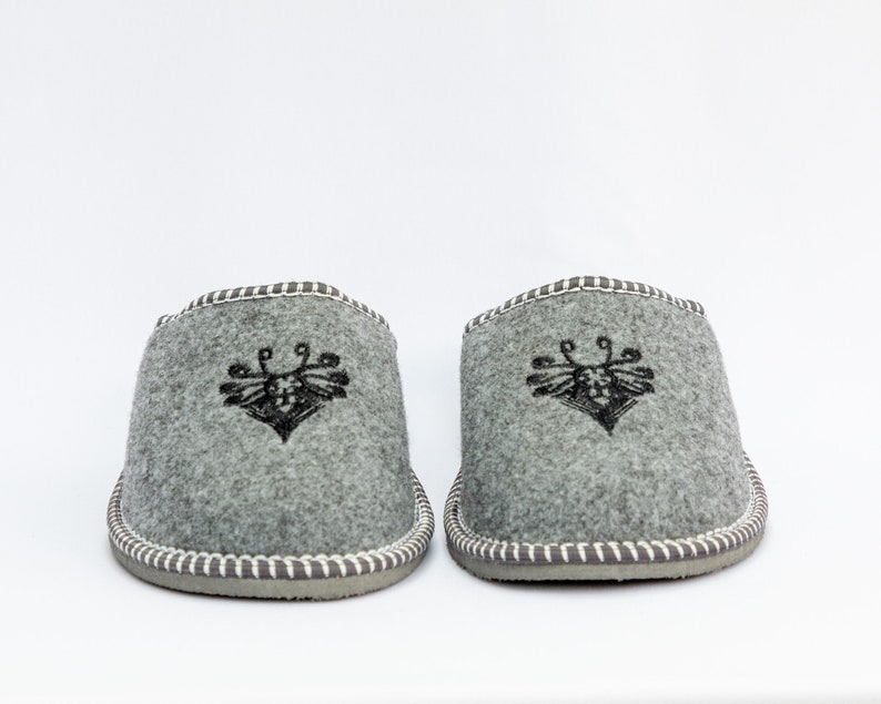 May include: A pair of gray wool slippers with a black embroidered bee design. The slippers have a white stitched trim around the edges.