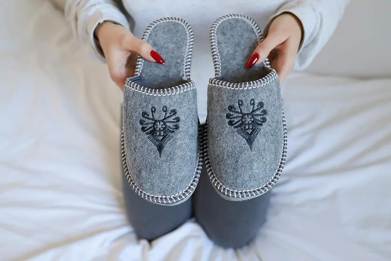 May include: A pair of gray felt slippers with a black embroidered design. The slippers have a white trim around the edges.