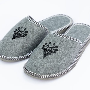 May include: A pair of gray felt slippers with black embroidered designs. The slippers have a white stitched trim around the edges.