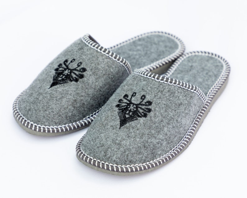 May include: A pair of gray felt slippers with black embroidered designs. The slippers have a white stitched trim around the edges.