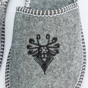 May include: Gray felt slippers with a black embroidered design on the top. The slippers have a white and black trim around the edge.