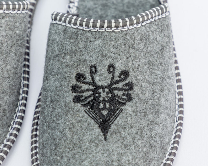 May include: Gray felt slippers with a black embroidered design on the top. The slippers have a white and black trim around the edge.
