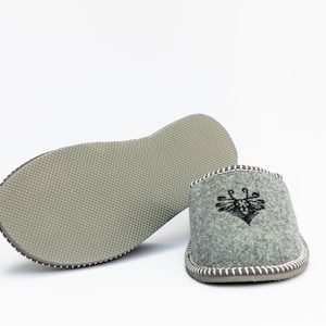 May include: A pair of gray felt slippers with a black embroidered design on the top. The slippers have a gray rubber sole.