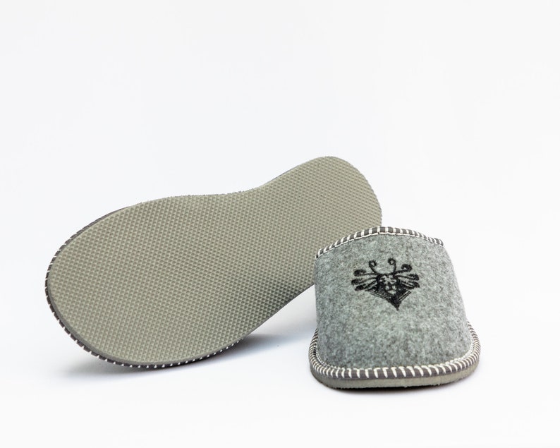May include: A pair of gray felt slippers with a black embroidered design on the top. The slippers have a gray rubber sole.