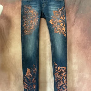 May include: Blue denim jeans with copper-coloured floral designs on the front and lower legs. The jeans are hanging on a black hanger. The floral patterns are symmetrical and detailed.