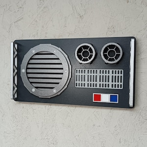 May include: A black and silver wall decoration featuring a circular vent, two circular designs, and a rectangular panel with vertical lines. It also has a small section with red, white, and blue rectangles.
