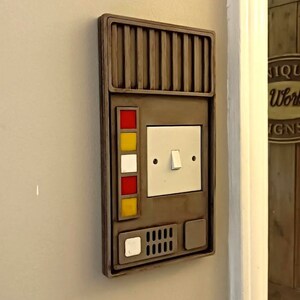 May include: A brown wall-mounted light switch with a futuristic design. It features a white light switch, four coloured buttons, and a small speaker with a white button.
