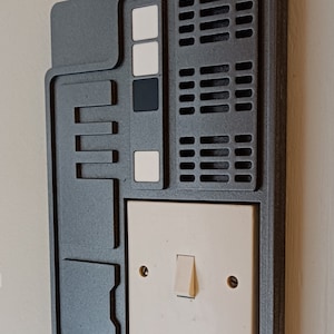 May include: A grey wall plate with a retro-futuristic design, featuring a light switch, small square buttons, and rectangular vents. The plate is mounted on a light-coloured wall. The design has a sci-fi aesthetic.