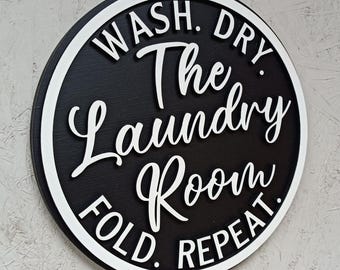 May include: A round black and white wooden sign with the text "Wash. Dry. The Laundry Room. Fold. Repeat." in white lettering.