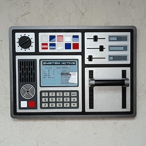 May include: A retro-style control panel with a gray frame. It features dials, buttons, sliders, and a keypad. The panel includes a screen displaying "SYSTEM ACTIVE" and other text. The color scheme is primarily gray, black, red, and blue.