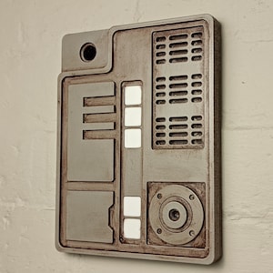May include: A beige wall mounted intercom with a rectangular shape and a circular button. The intercom has a series of rectangular buttons and a circular button with a small hole in the centre.