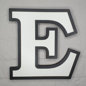 May include: A large, white, uppercase letter "E" with a black outline. The letter is made of wood and has a slightly distressed finish.